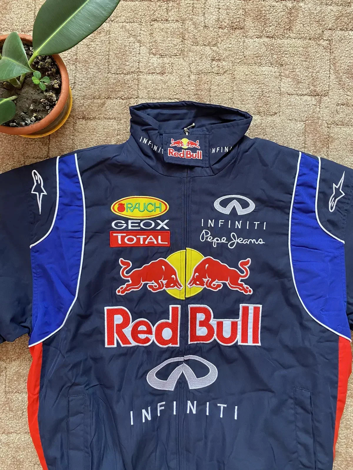 Navy blue Red Bull racing jacket with sponsor logos and bold embroidery, laid on tan carpet.